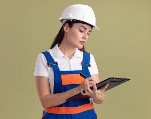 confident young builder woman in uniform writing something on clipboard isolated on olive green background