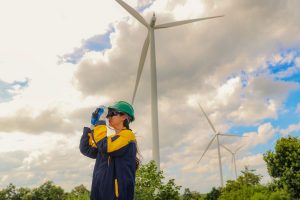 Female geographer in green safety helmet using binoculars surveying looking from distance working field in the wind farm clean energy project area the open terrain is suitable using wind energy